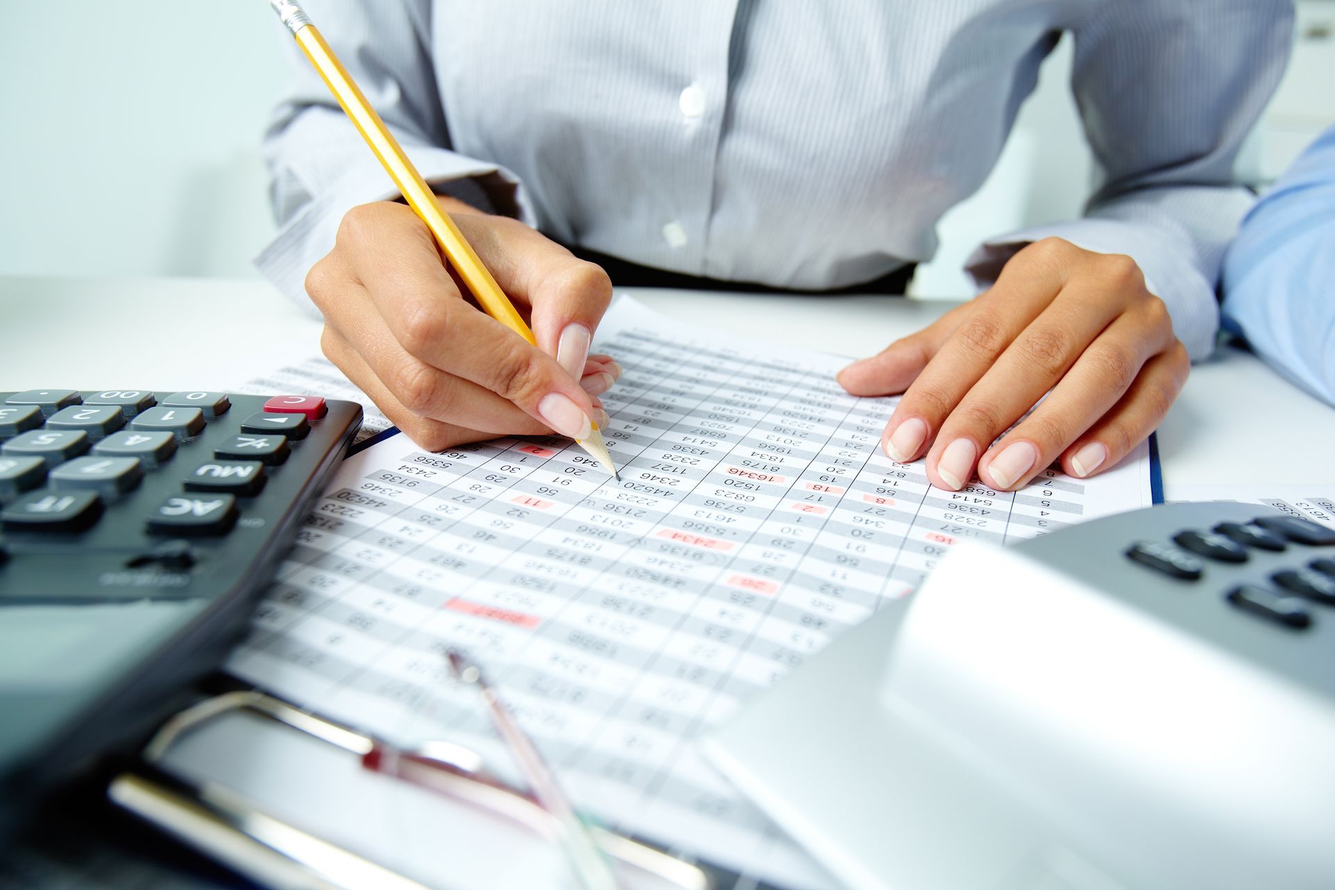Person in office, writing on a document with a pencil; calculator, phone, and glasses visible.